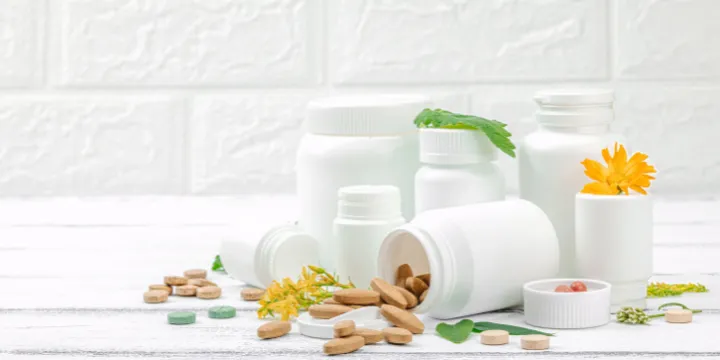 White supplement containers on a kitchen counter with colorful herbs and botanical ingredients arranged nearby in natural light