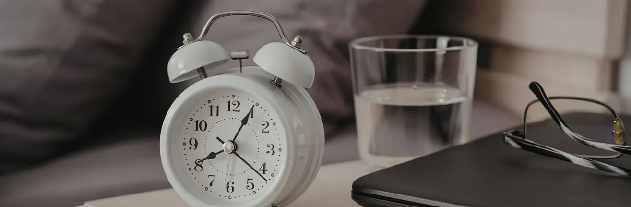 Bedside table with an alarm clock, glass of water, glasses, and a closed laptop next to the bed in soft natural light
