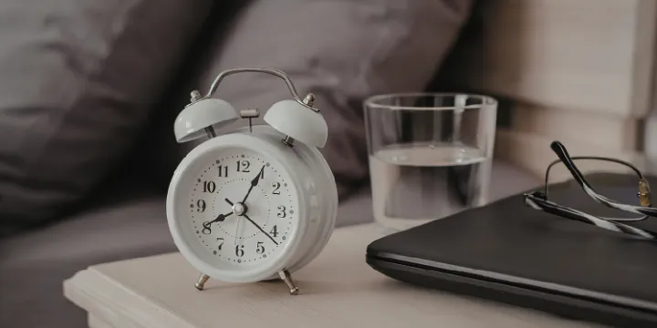 Bedside table with an alarm clock, glass of water, glasses, and a closed laptop next to the bed in soft natural light