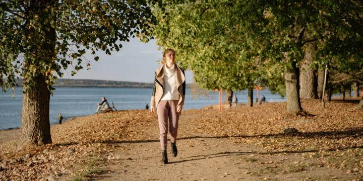 Woman walking along a long path beside a lake with trees and fallen leaves in natural light