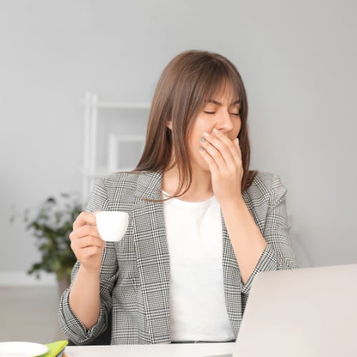 a sleep deprived woman at work yawning and drinking a cup of coffee to stay awake