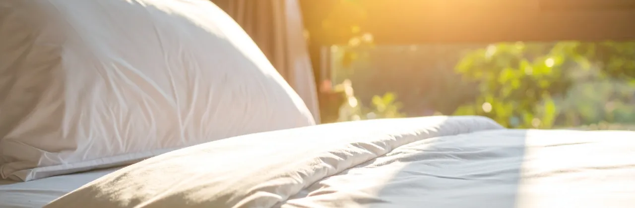 Morning sunlight filtering through bedroom curtains, softly illuminating a bed and pillow in a calm, quiet space