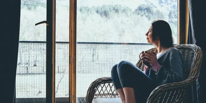 Woman sitting comfortably in a chair at home with a cup of tea, looking out a window in natural light