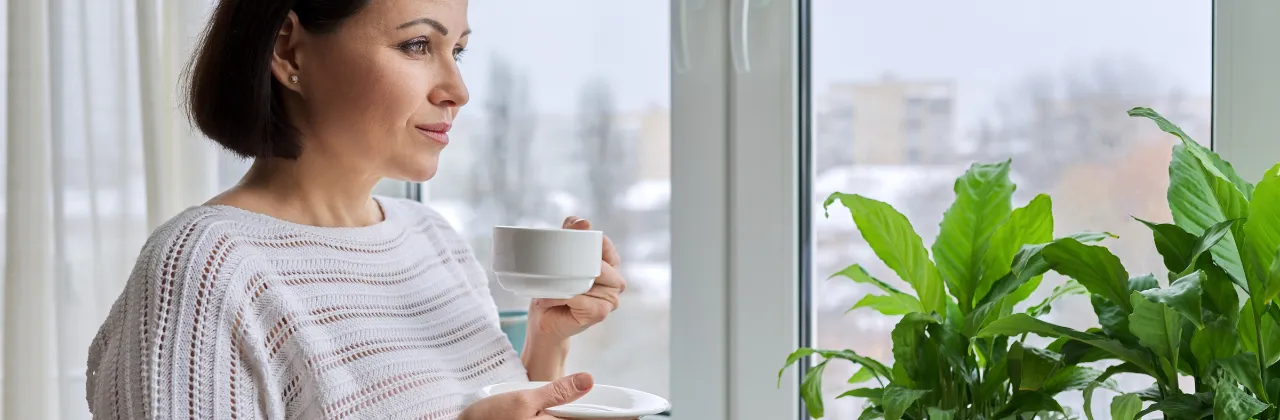 Middle-aged woman holding a cup of coffee, gazing out a window toward a muted city view with indoor plants nearby