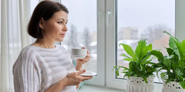 Middle-aged woman holding a cup of coffee, gazing out a window toward a muted city view with indoor plants nearby